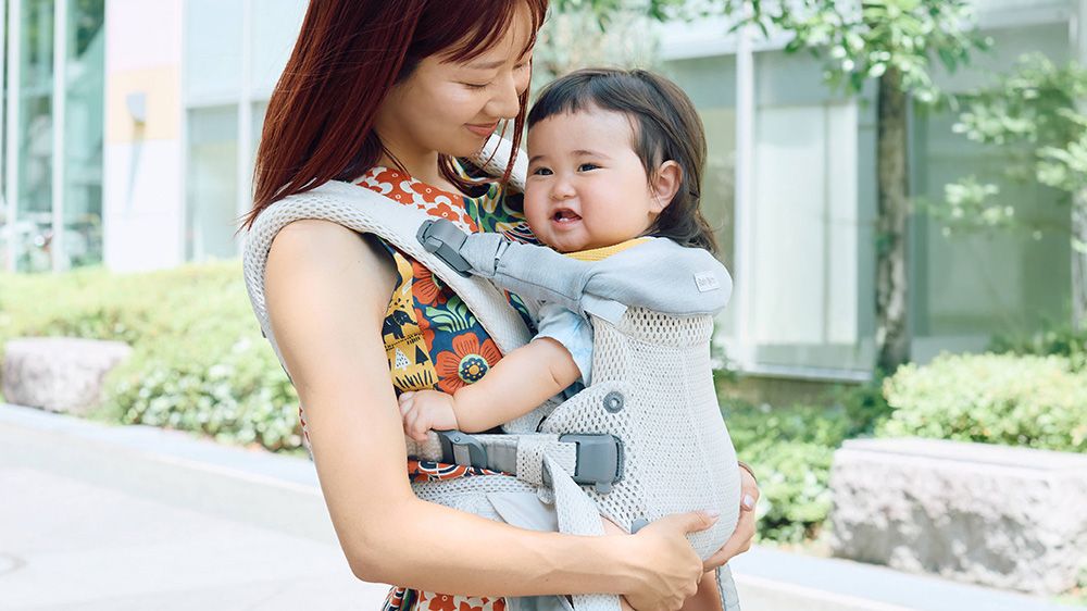 A woman smiles at a baby in a gray carrier outdoors. The baby is also smiling, and they are surrounded by greenery and modern buildings.