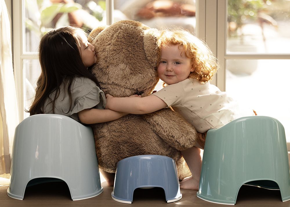 Two children sit on potties, hugging a large teddy bear by a sunlit window.