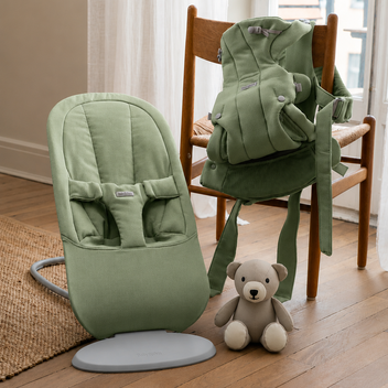 Green baby bouncer and carrier on a wooden chair next to a plush teddy bear on a wooden floor by a curtain.