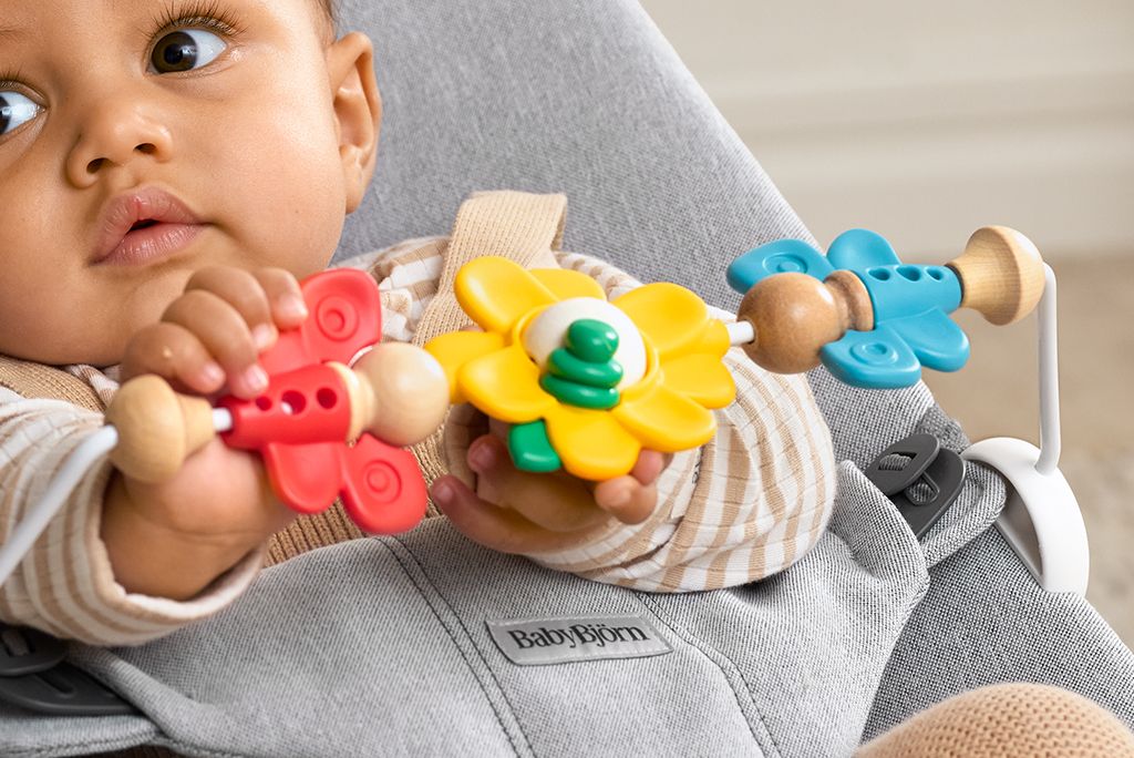 Baby in a striped outfit plays with colorful flower-shaped toys on a gray BabyBjörn bouncer.