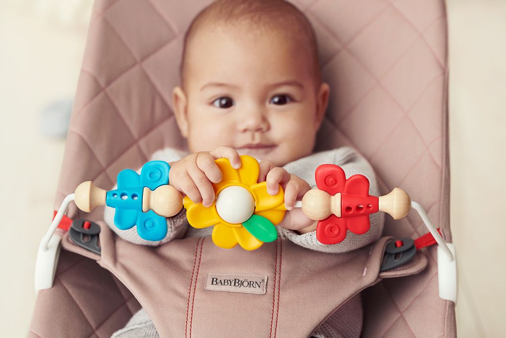 Baby sitting in a stroller, holding a colorful toy with blue and red flowers, wearing a light-colored outfit.