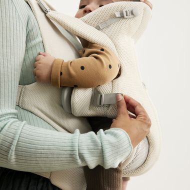 Person wearing a light green sweater holds a baby in a white carrier. The baby is dressed in a brown polka dot outfit and appears snug.