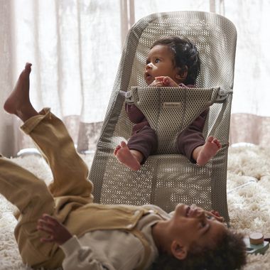 A baby in a mesh baby bouncer looks up curiously as an older child wearing overalls lies playfully beside them on a fluffy rug.