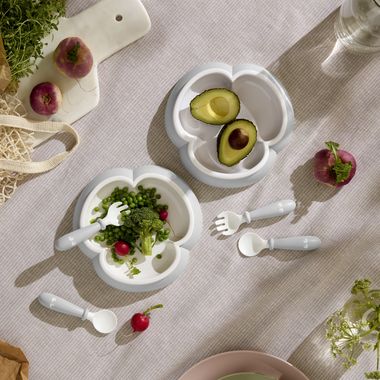 Two white toddler plates with utensils, one holding avocado halves and a radish, the other with peas, cilantro, and radishes on a striped tablecloth.