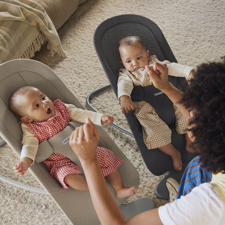 Two babies in checkered outfits sit in baby bouncers, reaching towards an adult sitting nearby on a carpeted floor.