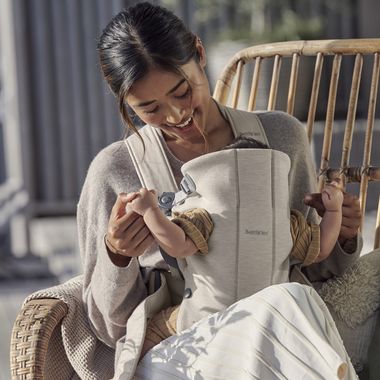 A woman sits on a wicker chair, smiling at a baby in a beige carrier. The baby is wearing a brown outfit, and they are outdoors.