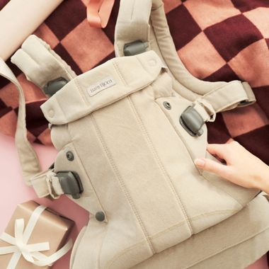 A beige baby carrier on a checkered blanket with a hand adjusting it, alongside a wrapped gift with a white ribbon.
