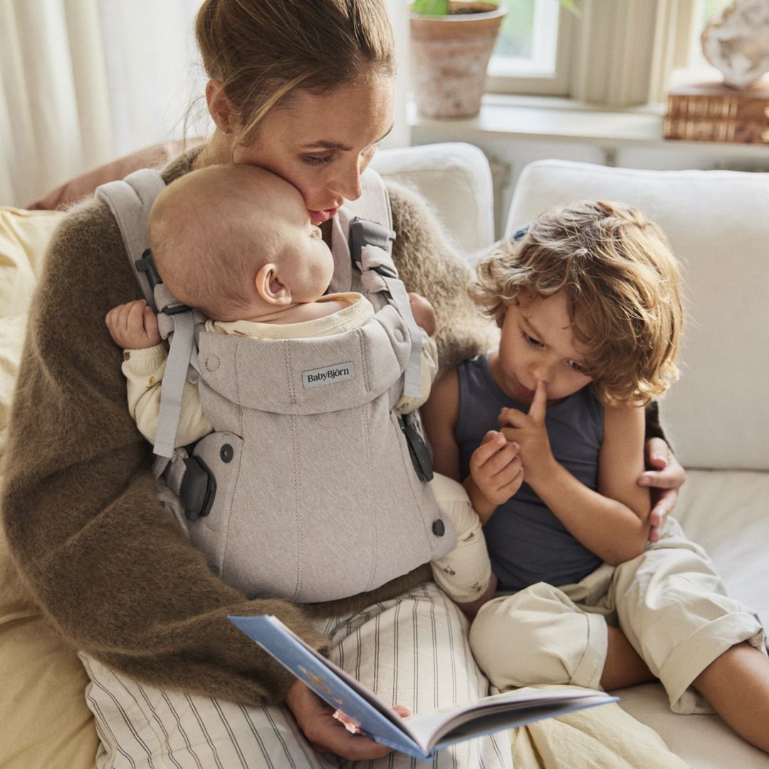 Mother holding baby in carrier, reading book with young child on sofa. Baby and child focused on the book, creating an intimate family moment.