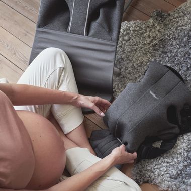 Pregnant person sitting on a wooden floor near a soft rug, holding a gray baby carrier.