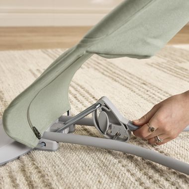 A person in green pants engages a latch mechanism on a gray folding metal frame on a textured beige rug.