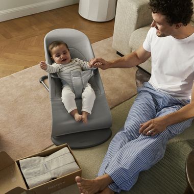 A man in casual clothes holds hands with a smiling baby in a gray bouncer on a living room floor next to an open box.