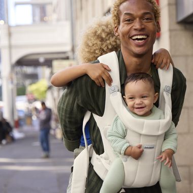 Man smiling, carrying a laughing baby in a front carrier and another child on his back, walking outdoors.