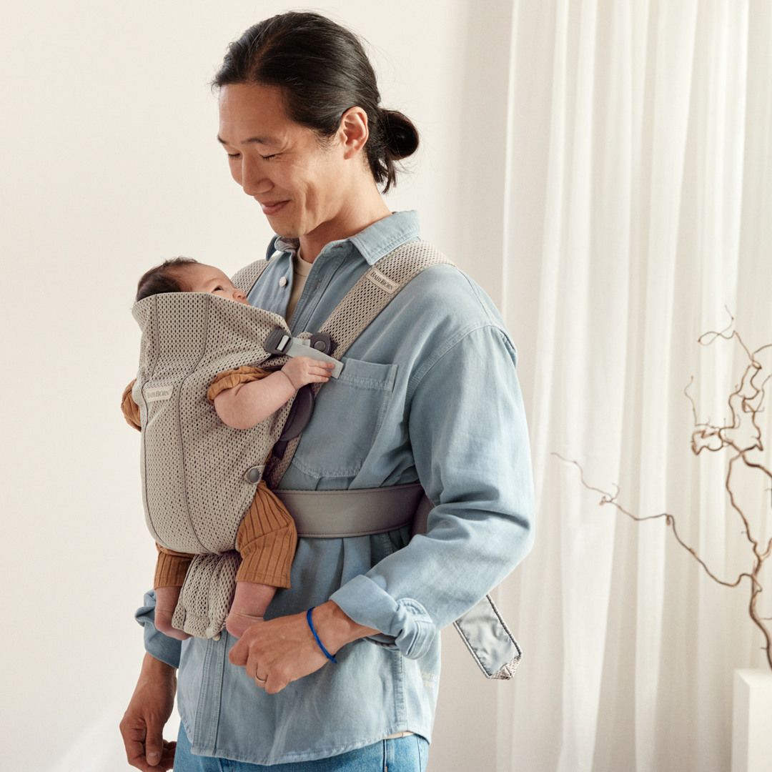 A person in a blue shirt smiles down at a baby in a beige carrier, standing in a softly lit room with white curtains and a decorative branch.