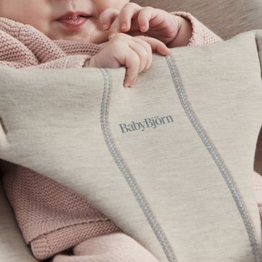 A baby wearing a pink knitted outfit is partially visible, resting in a beige BabyBjörn carrier, with fingers near the mouth.