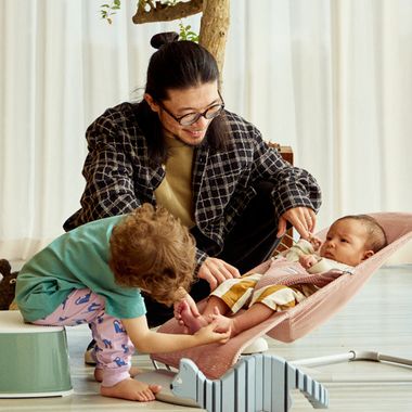 A smiling man watches a toddler gently touch a baby's feet in a pink mesh BabyBjörn bouncer bliss. The scene is warm and playful, with a cozy home atmosphere.