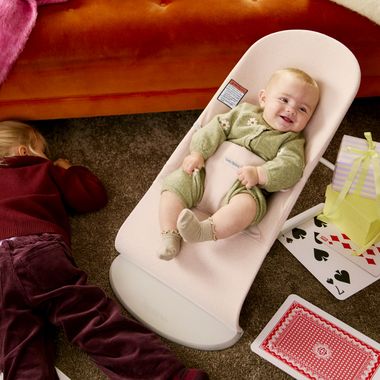 Smiling baby in a pale pink bouncer on a carpeted floor, an older child lying nearby, oversized playing cards and a small wrapped gift beside them.