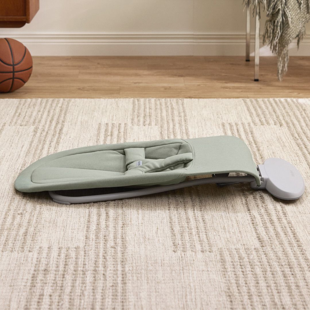 A portable green baby bouncer on a beige patterned rug, with a basketball and furniture in the background.