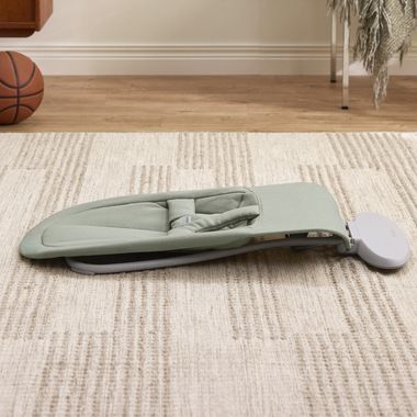 A portable green baby bouncer on a beige patterned rug, with a basketball and furniture in the background.