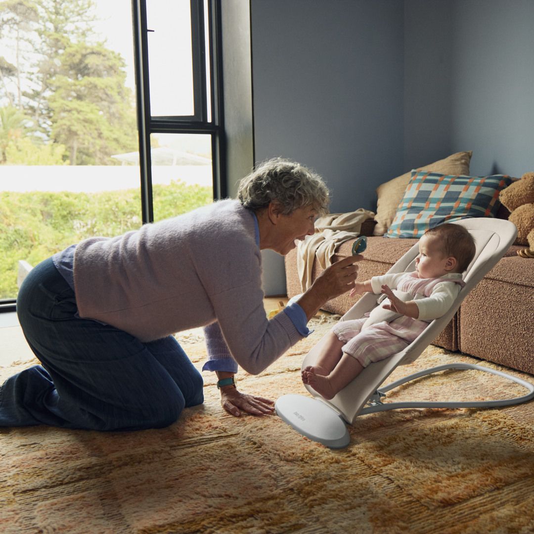 Older person kneeling and playing with a baby in a bouncer chair inside a cozy living room near a window.