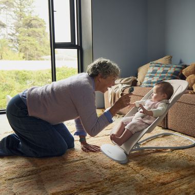 Older person kneeling and playing with a baby in a bouncer chair inside a cozy living room near a window.