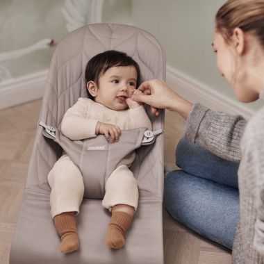 Baby seated in a beige bouncer, wearing a cream outfit and brown socks, playfully touching an adult's hand.