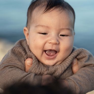 A joyful baby in a brown sweater is held up by hands, smiling with eyes closed near a beach setting.