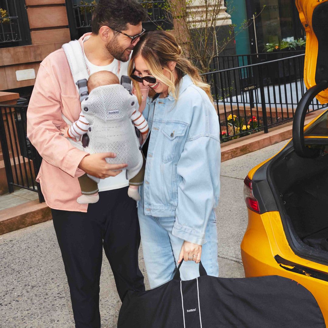 A smiling couple stands near an open car trunk, with a baby in a carrier. The woman holds a large bag.