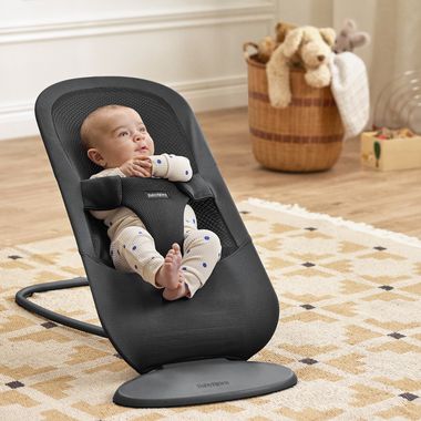A baby in a polka dot onesie sits in a black baby bouncer on a patterned rug, with a wicker basket and toys in the background.