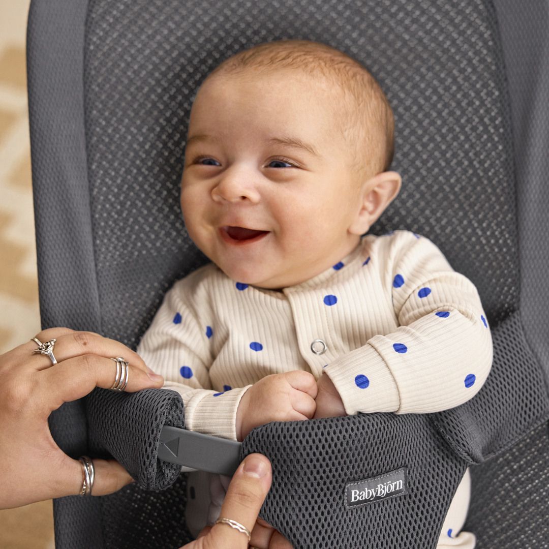 A smiling baby in a white outfit with blue polka dots sits in a gray BabyBjörn bouncer while being held by an adult's hands.