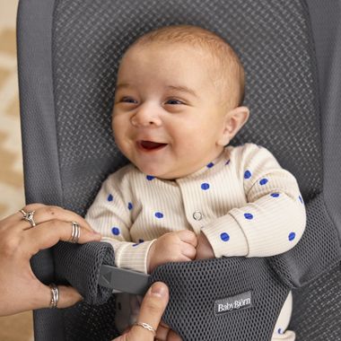 A smiling baby in a white outfit with blue polka dots sits in a gray BabyBjörn bouncer while being held by an adult's hands.