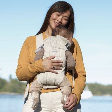 A smiling woman carries a sleeping baby in a beige baby carrier outdoors, with a blurred background of water and trees.