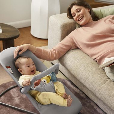 A smiling woman relaxes on a sofa, gently touching a baby in a gray bouncer. The baby is engaged with a colorful toy bar.