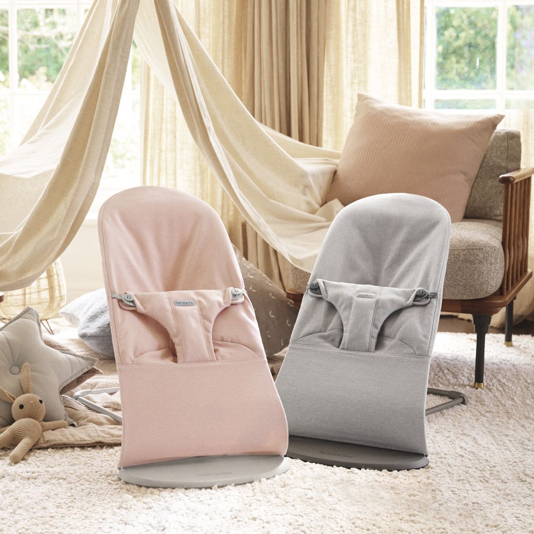 Two baby bouncers, one pink and one gray, placed in a cozy room with a beige tent, plush pillows, and soft rugs by a window.