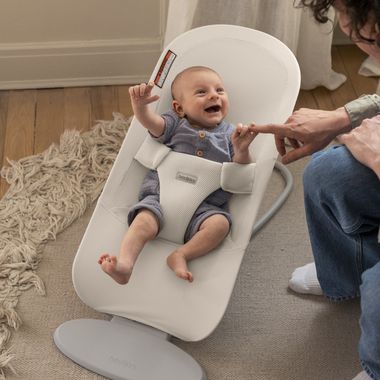 A smiling baby in a grey romper sits in a white bouncer, reaching for an adult's fingertip. The scene is warm and playful.