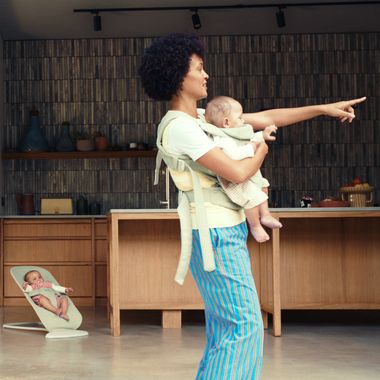 A person wearing a baby carrier points forward while holding a baby. Another baby is in a bouncer nearby in a modern kitchen.