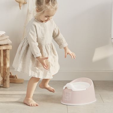 Young girl in a beige dress, barefoot, playfully stepping near a pink smart potty from BabyBjörn in a bright, minimalist room.