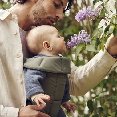 A man with a beard holds a baby in a green carrier, both closely observing purple flowers on a leafy bush.