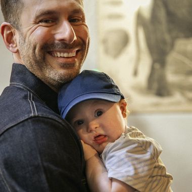 A smiling man holding a baby wearing a blue cap. The baby looks at the camera while resting on his shoulder.