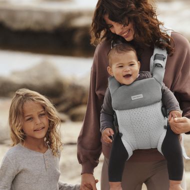 A woman smiles while holding hands with a young child and carrying a baby in a gray carrier at the beach.