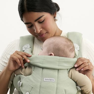 A woman gently cradles a baby in a soft green baby carrier, looking down affectionately at the child nestled against her chest.