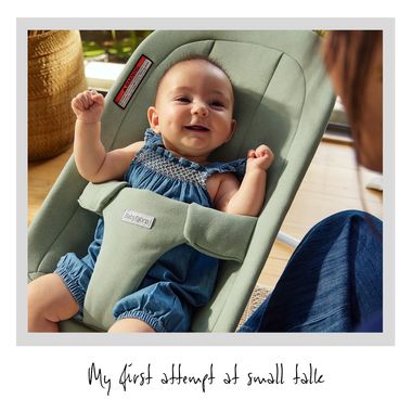 A smiling baby in a blue romper sits in a green bouncer. A person is interacting with the baby, creating a warm, playful scene.