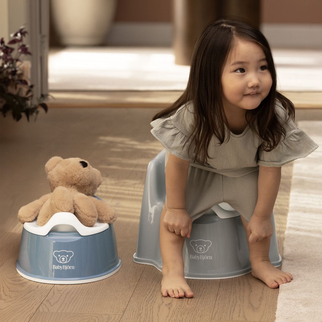 Young girl smiling while sitting on a blue potty chair, next to a teddy bear on a similar chair. Bright indoor setting.