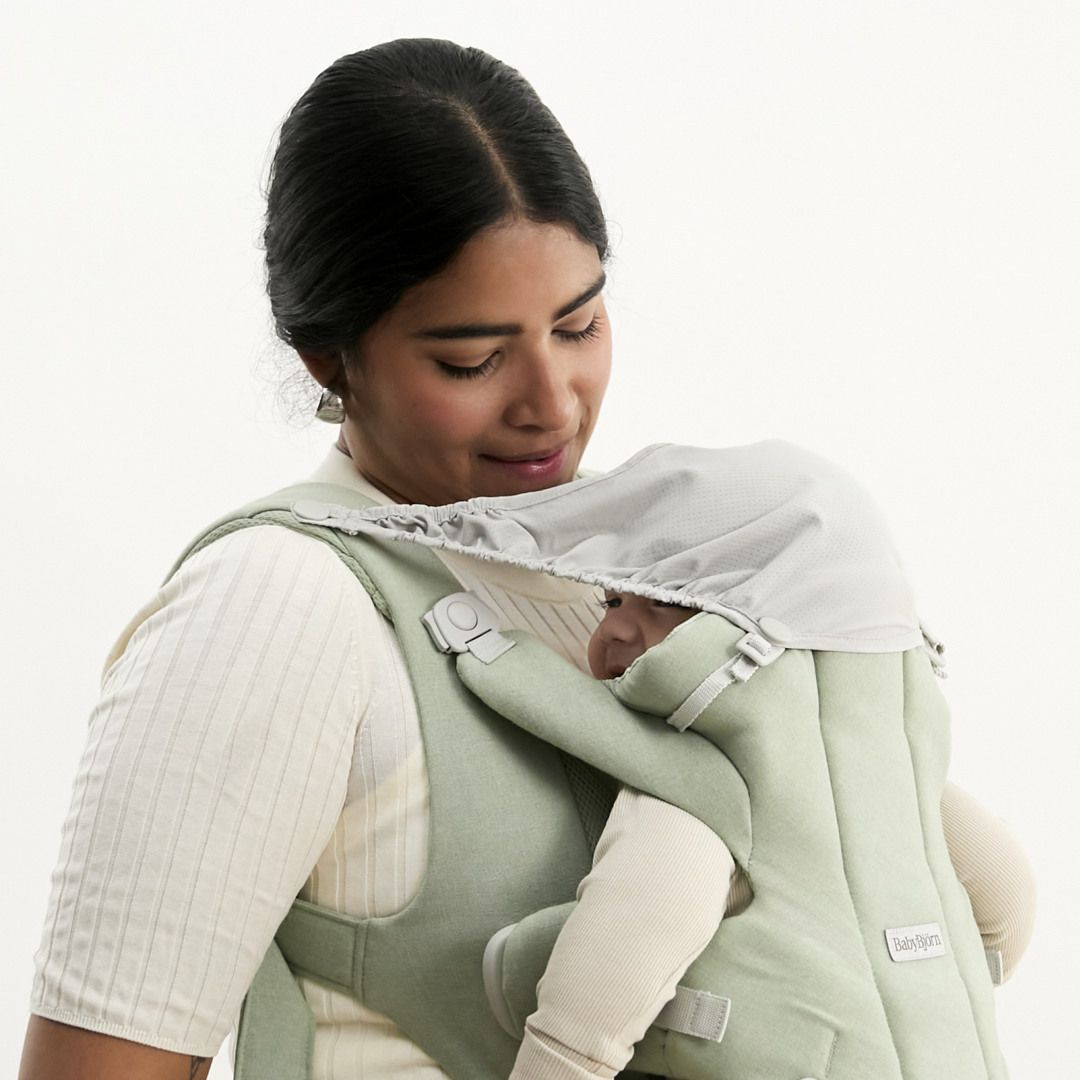 A woman gently cradles a baby in a soft green baby carrier, looking down affectionately. The baby is partially covered by a shade.
