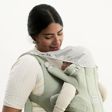 A woman gently cradles a baby in a soft green baby carrier, looking down affectionately. The baby is partially covered by a shade.