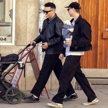 Two people walk down a city street, one pushing a stroller and the other carrying a baby in a sling, both wearing casual outfits.