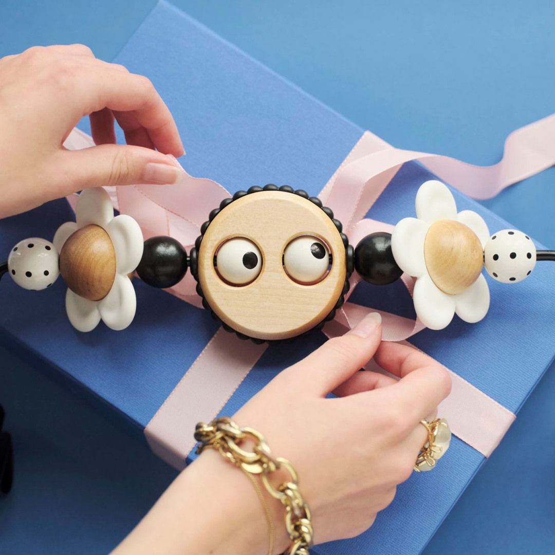 Hands holding a toy with googly eyes and flower designs over a blue gift box with a pink ribbon.