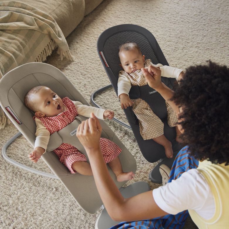 A caregiver plays with two babies seated in bouncers on a carpeted floor, both wearing checkered outfits.
