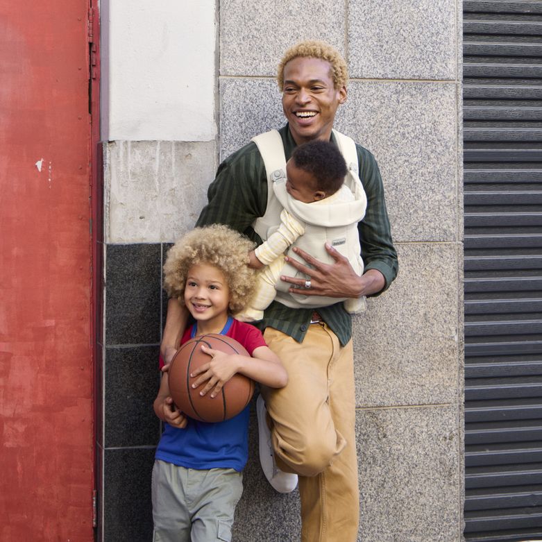 A smiling man leans against a wall with a baby in a carrier and a child holding a basketball.