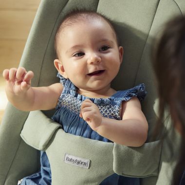 A smiling baby in a blue dress sits in a green baby bouncer, reaching out with one hand.