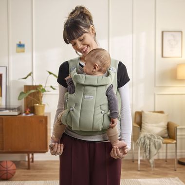 A woman smiles at a baby in a green carrier at home, with a wooden cabinet, plant, and basketball in the background.